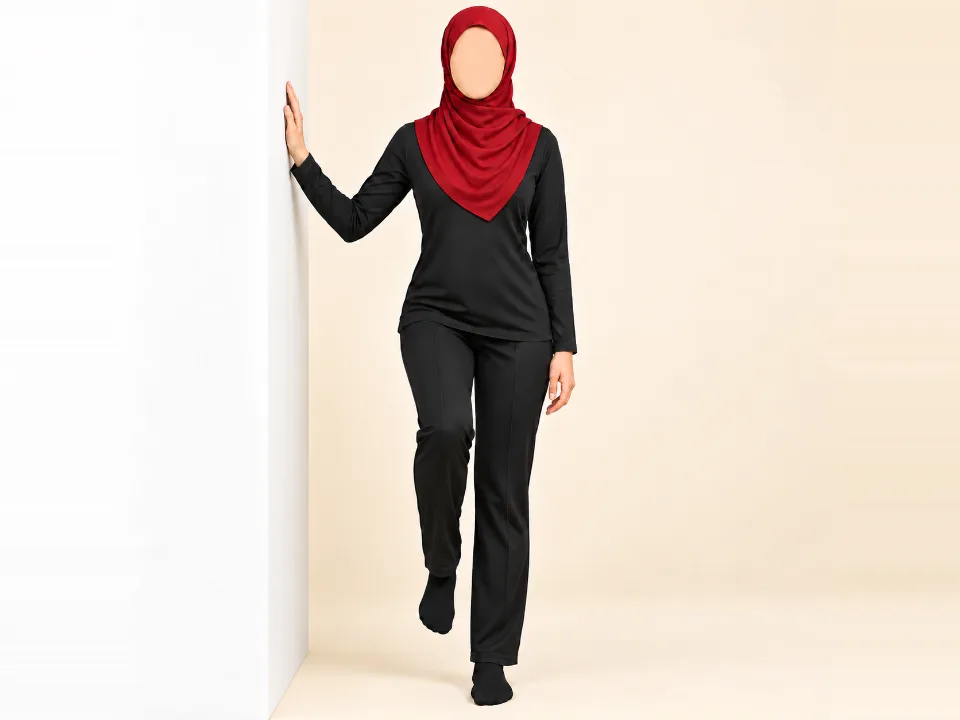 Woman in a red hijab standing on one leg beside a wall with her fingertips on it and her hips level.