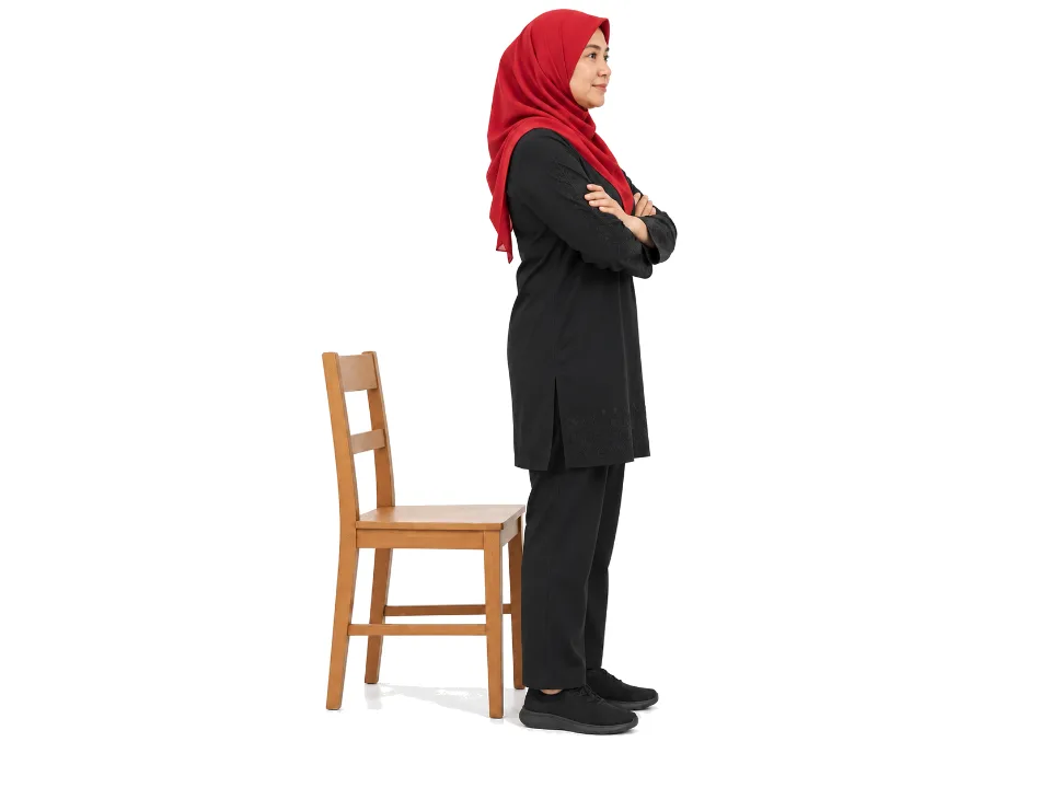 Illustrative image, not a real patient. A woman in a red hijab stands upright in front of a wooden chair with her arms crossed over her chest.