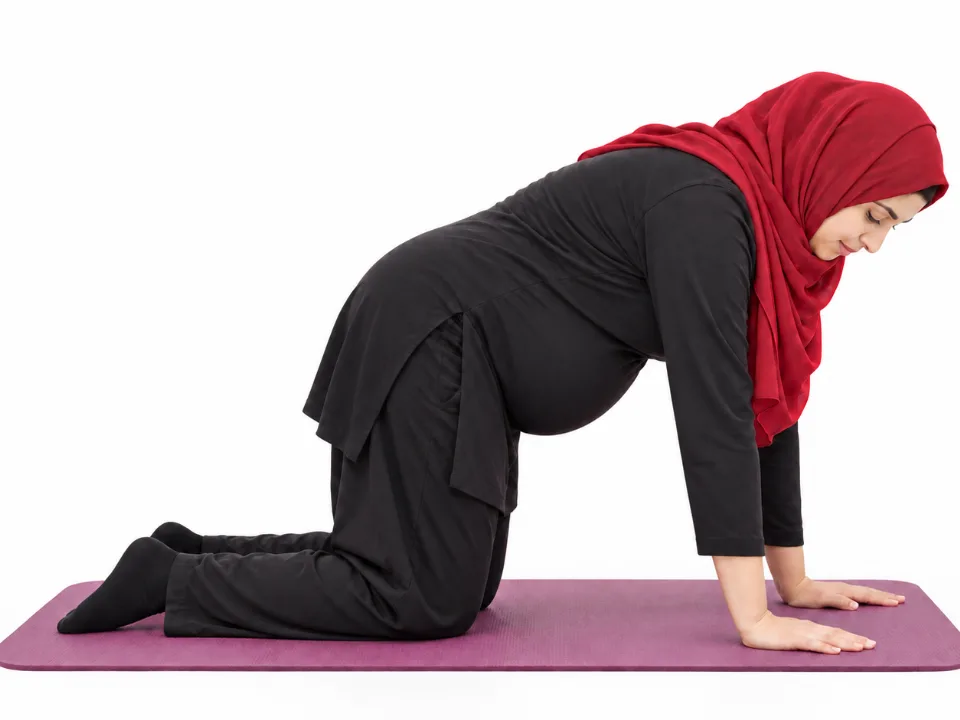 Illustrative image, not a real patient. A pregnant woman in a red hijab kneels on all fours on a mat with her lower back gently rounded.